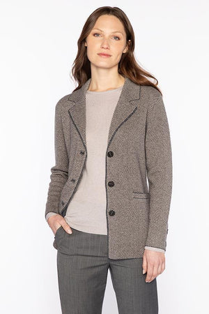 A woman with wavy brown hair wears the KINROSS CASHMERE Herringbone Notch Collar Knit Cardigan under a gray blazer with matching pants, standing against a white background with one hand in her pocket.