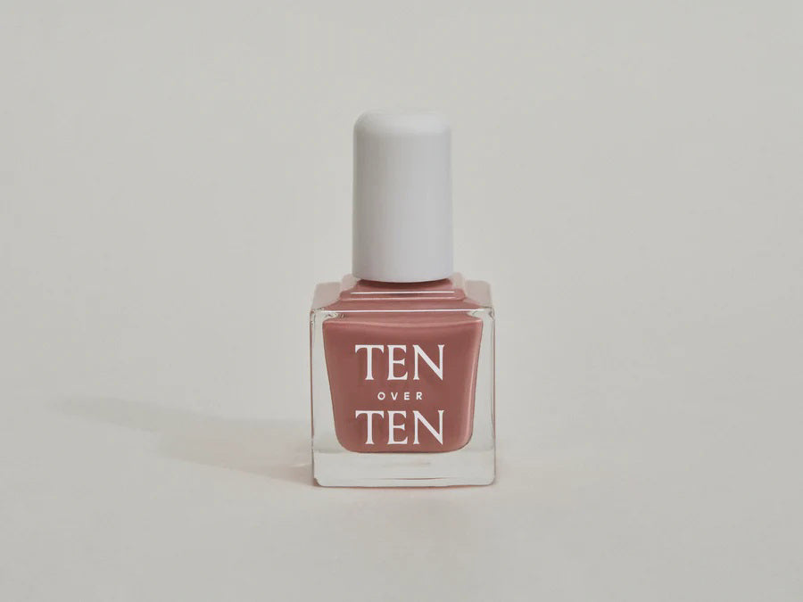 A glass bottle of TENOVERTEN - NAIL POLISH from the brand TEN OVER TEN, featuring a white cap and a dusty pink nail color. The front label displays "TEN OVER TEN" in white uppercase letters. This vegan, eight-free formula ensures both beauty and peace of mind. The background is plain and light-colored.