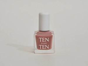 A glass bottle of TENOVERTEN - NAIL POLISH from the brand TEN OVER TEN, featuring a white cap and a dusty pink nail color. The front label displays "TEN OVER TEN" in white uppercase letters. This vegan, eight-free formula ensures both beauty and peace of mind. The background is plain and light-colored.