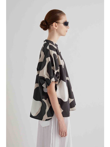 A person with brown hair in a low bun is shown from the back, wearing the FRANK AND EILEEN - MOONBEAM TOP by MEIMEIJ—an abstract print top with loose, short sleeves in black, white, and beige—paired over a white pleated skirt.