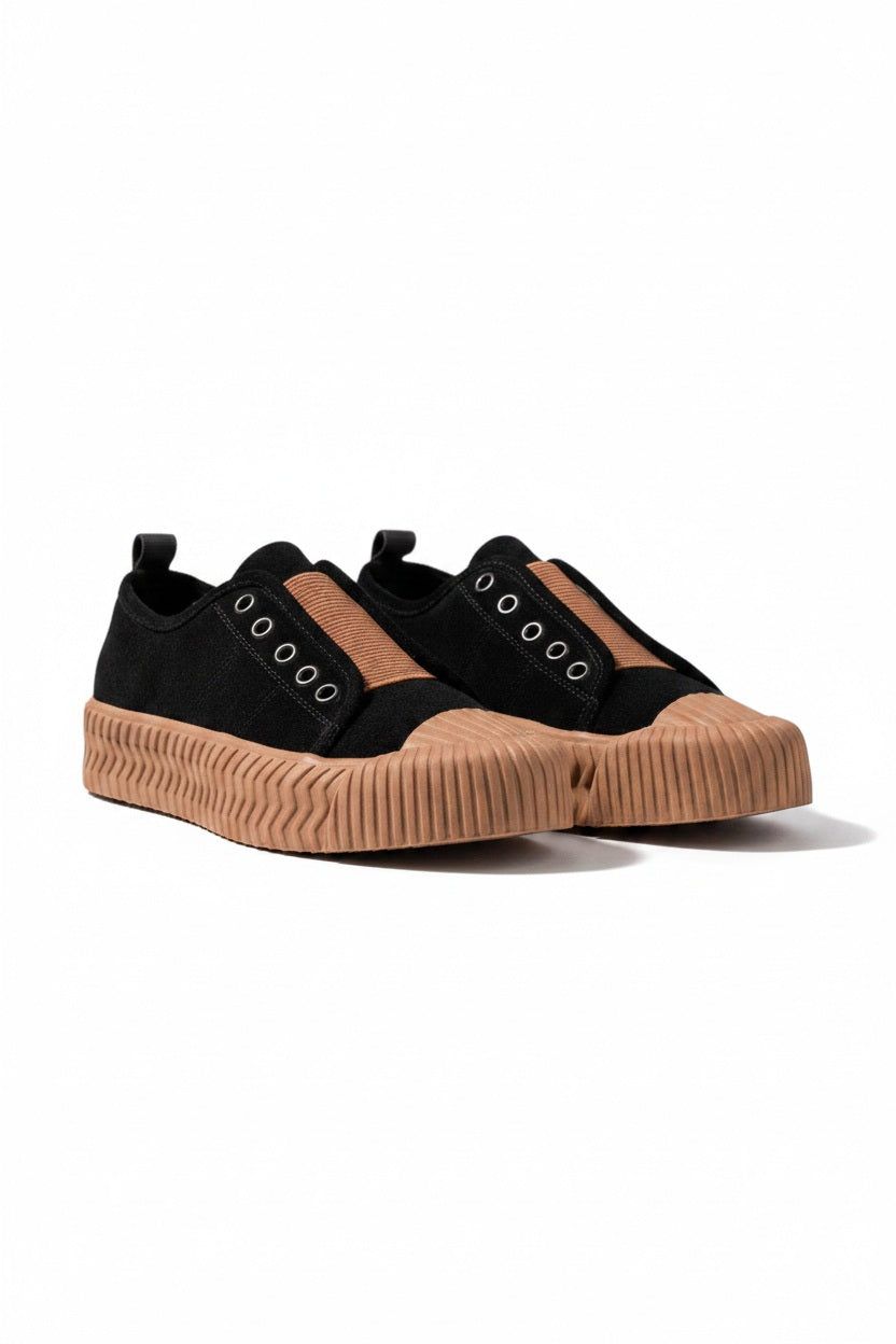 The DANIELLA LEHAVI - OSAKA SNEAKER in black, designed for daily wear, features thick tan rubber soles and a brown fabric strip, shown on a white textured pedestal with a neutral background.