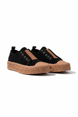 The DANIELLA LEHAVI - OSAKA SNEAKER in black, designed for daily wear, features thick tan rubber soles and a brown fabric strip, shown on a white textured pedestal with a neutral background.