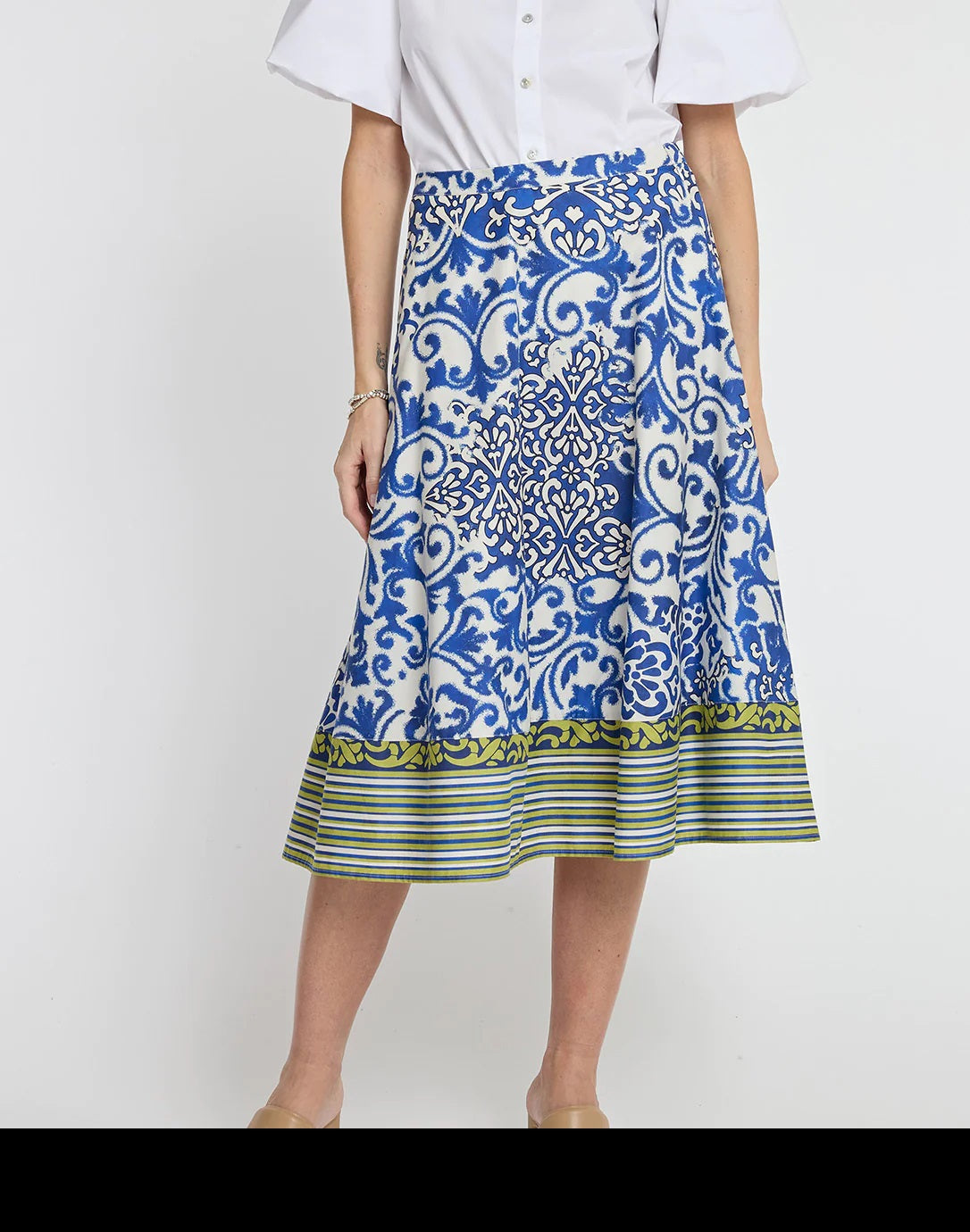 A person, whose head and feet are not visible, stands against a plain background wearing a white blouse and the HINSON WU - GLORIA SKIRT IN CERAMIC TILE PRINT midi skirt by HINSON WU, featuring a blue and white ceramic tile print with a green striped hem.