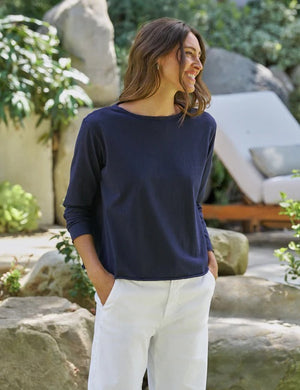 A woman with wavy brown hair stands outdoors among greenery and rocks, facing away but looking over her shoulder with a smile. She wears the FRANK & EILEEN Ella Long Sleeve Boatneck Tee in Classic Navy, paired with white pants.