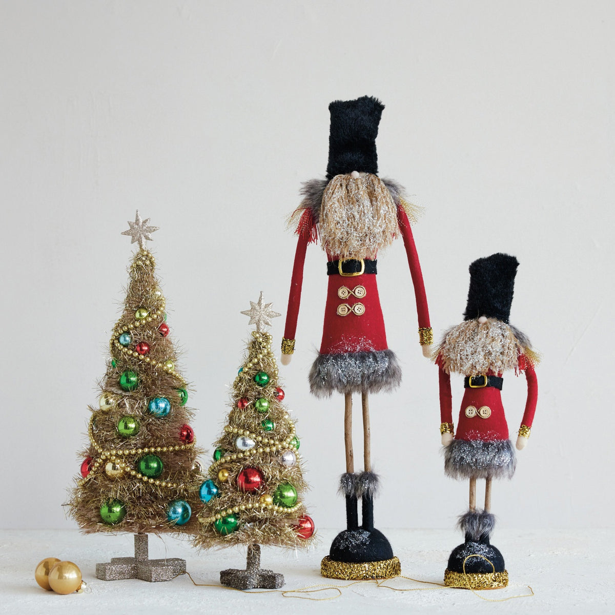 Two decorated gold tinsel Christmas trees stand next to two FABRIC SOLDIER WITH GLITTER, FAUX FUR AND TWIG LEGS figurines.