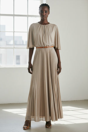A woman poses against a plain background wearing the MOMONI - PARADISU DRESS by MOMONI, featuring light beige lurex jersey, kimono sleeves, and a pleated skirt. She styles it with braided hair, earrings, and open-toed shoes.