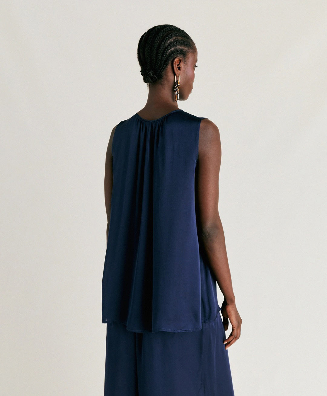 A person stands against a plain background wearing the MOMONI - CRIS SLEEVELESS TOP, a loose-fitting, round neck top in an elegant navy blue. They are also dressed in matching wide-leg pants crafted from pure silk, creating a sophisticated and flowing look. The individual has short, styled hair and is glancing downward with one hand touching their ear.