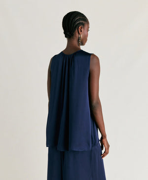 A person stands against a plain background wearing the MOMONI - CRIS SLEEVELESS TOP, a loose-fitting, round neck top in an elegant navy blue. They are also dressed in matching wide-leg pants crafted from pure silk, creating a sophisticated and flowing look. The individual has short, styled hair and is glancing downward with one hand touching their ear.
