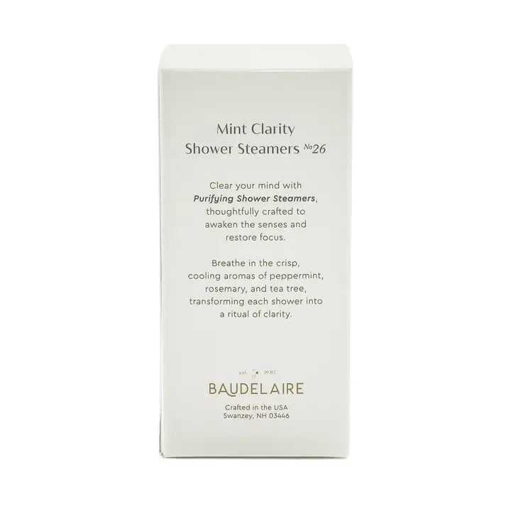 A white box labeled “BAUDELAIRE - PURIFY MINT CLARITY SHOWER STEAMER” contains light blue shower steamers stacked neatly inside, crafted for aromatherapy by BAUDELAIRE. The steamers, infused with essential oil blends, are visible through a clear window, and “Mint Clarity Shower Steamers No. 26” appears at the bottom of the box.