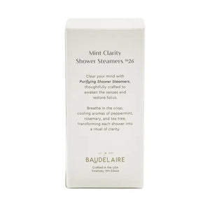 A white box labeled “BAUDELAIRE - PURIFY MINT CLARITY SHOWER STEAMER” contains light blue shower steamers stacked neatly inside, crafted for aromatherapy by BAUDELAIRE. The steamers, infused with essential oil blends, are visible through a clear window, and “Mint Clarity Shower Steamers No. 26” appears at the bottom of the box.