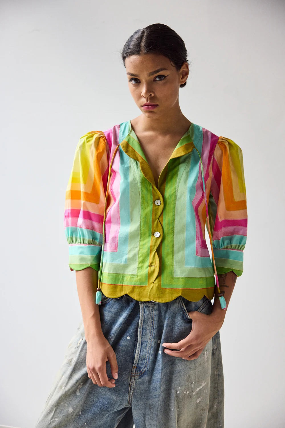 A person wears the HOPE FOR FLOWERS - SCALLOPED SHIRT IN CONCENTRIC SQUARES, featuring colorful geometric stripes and scalloped edges, paired with light blue jeans. The image highlights the torso.