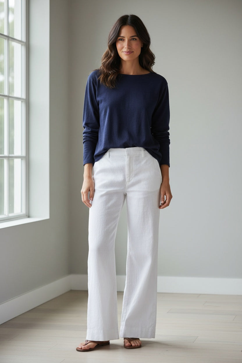 A woman stands outdoors, smiling with her hands in her pockets. She is wearing the FRANK &amp; EILEEN Ella Long Sleeve Boatneck Tee in Classic Navy, paired with white pants. The background features rocks, greenery, and an outdoor lounge chair.