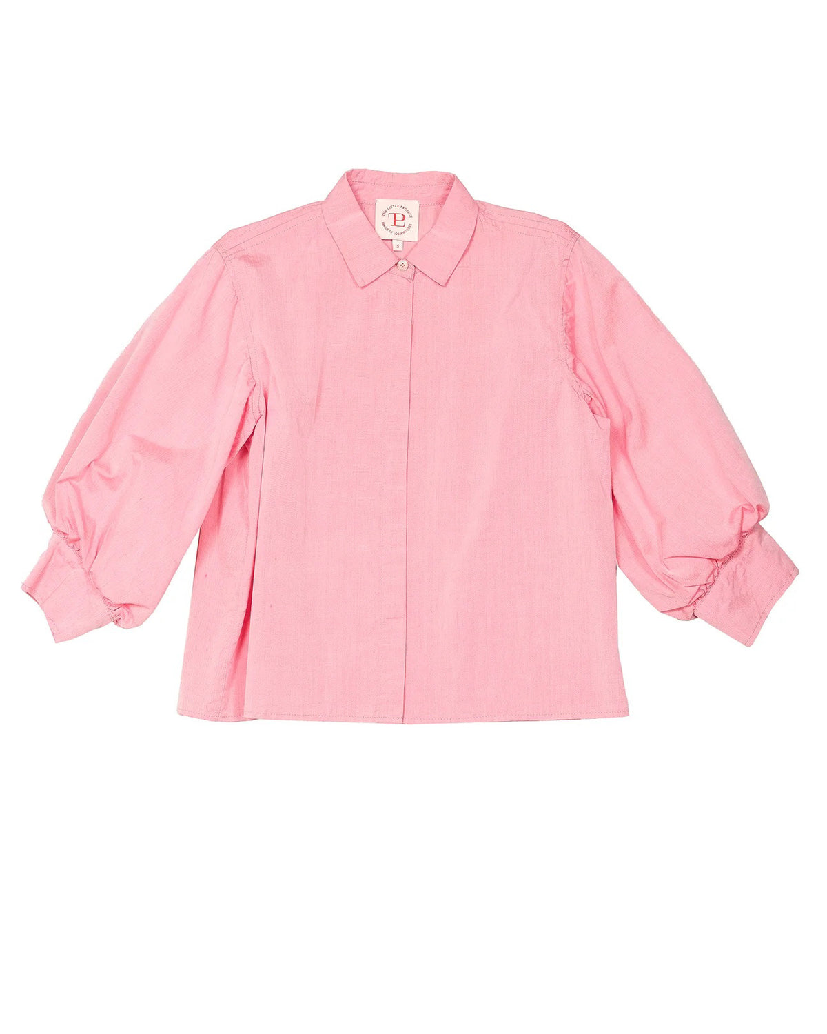 A pink button-front shirt, THE LITTLE PROJECT&#39;s ESME SHIRT IN STRAWBERRY, is displayed flat on a white surface. It has a straight cut with one visible button near the collar and a &quot;100% Linen&quot; tag featuring the brand logo and size label.