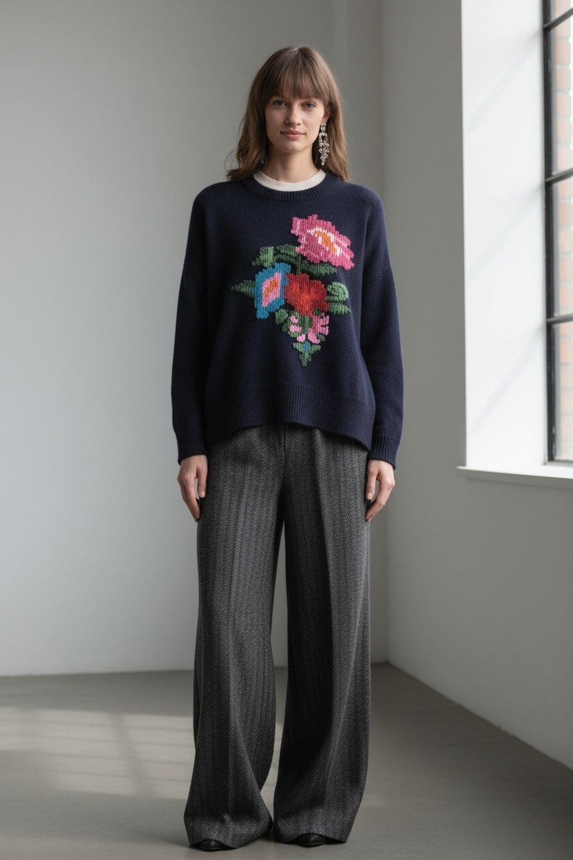 A person stands indoors by a window, wearing the FLOOR - NAVY WOOL SWEATER from FLOOR, which features a large floral motif, paired with loose, wide-leg gray pants. They have straight brown hair and are looking at the camera with a neutral expression.