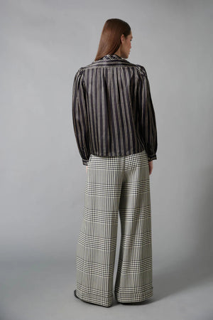 A woman with straight brown hair faces away in a HOPE FOR FLOWERS Tie Neck Blouse—black and white, vertically striped Tencel with puffed sleeves—paired with wide-leg plaid trousers. The plain gray background reflects sustainable style.