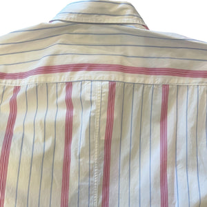 A man in a FRANK & EILEEN Finbar Men's Button Up Shirt in hot pink and blue stripe, blue jeans, and white sneakers walks confidently down a cobblestone street lined with stone buildings and potted plants on a sunny day.