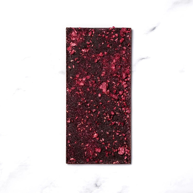 A rectangular chocolate bar with a pinkish wrapper adorned with minimalist illustrations of raspberries and abstract shapes. The packaging reads "RITUAL CHOCOLATE - THE APRES CHOCOLATE BAR 70%, Sparkling Wine Infused Chocolate with Dried Raspberries," 2.12 oz (60g).