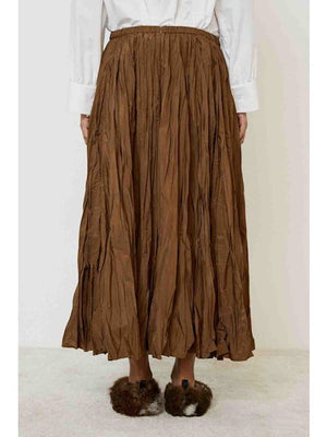 A person wearing the MEIMEIJ CRINKLE SKIRT stands against a plain light background. Only the lower half, featuring the brown midi skirt and brown fuzzy slippers, is visible.