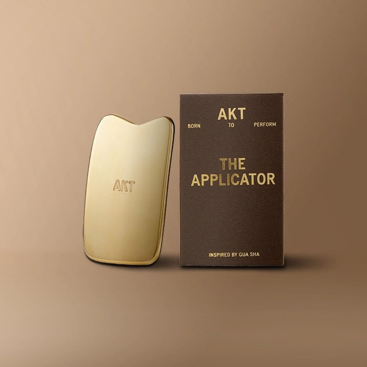 A gold massage tool labeled “AKT”, known as the AKT - THE APPLICATOR GUA SHA TOOL from AKT LONDON and inspired by traditional Gua Sha tools for lymphatic drainage, stands next to a brown box displaying the text “AKT TO PERFORM THE APPLICATOR INSPIRED BY GUA SHA” on a beige background.
