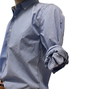 A man in a FRANK & EILEEN Finbar Men's Button Up Shirt in Blue Stripes, blue jeans, and white sneakers walks confidently down a sunny, tree-lined cobblestone street.