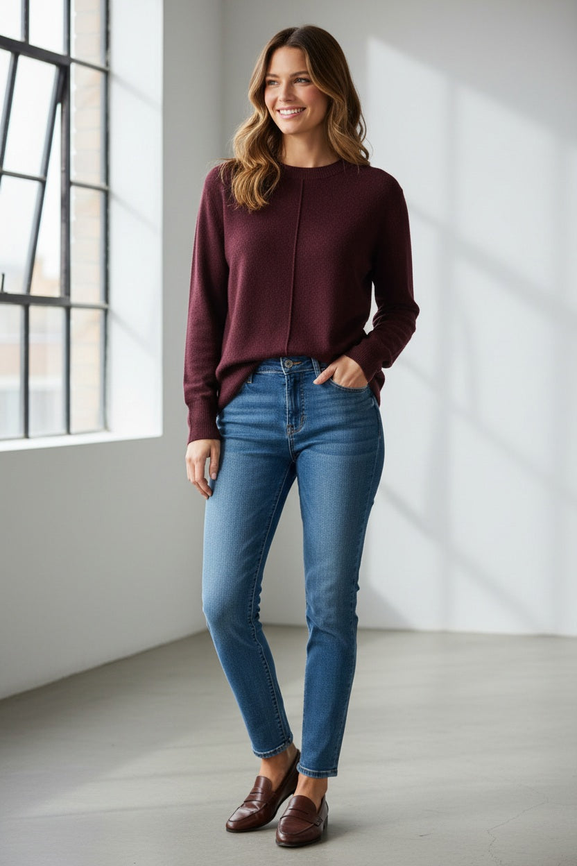 A person with long light brown hair wears the KINROSS CASHMERE Hi Low Seamed Crew Neck Cashmere Sweater in Cordovan—featuring a relaxed fit—paired with blue jeans, standing against a plain light background and facing forward with a neutral expression.