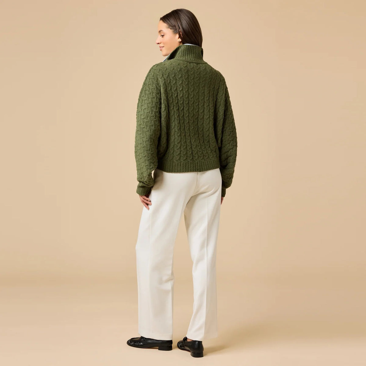 A woman wearing a cream-colored, plush SOFTIES - CABLE KNIT MARSHMALLOW FUNNEL NECK DOUBLE ZIP JACKET with a funnel neck and double zipper, paired with blue jeans.
