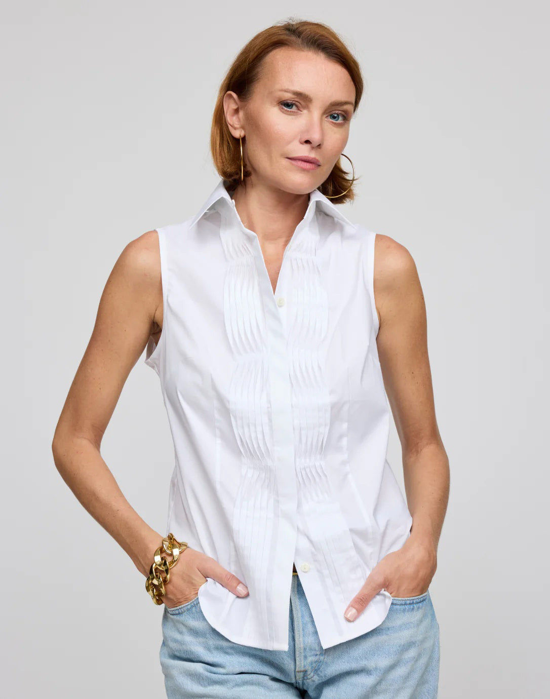 A woman with long hair wears the HINSON WU Lilith Sleeveless Shirt, made from stretch polished cotton with pleats and a beige belt, against a gray background. She exudes confidence as she gazes at the camera, highlighting the shirt&#39;s versatility.