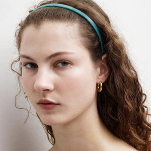 A young woman with wavy brown hair wears the MACHETE Ultralight Thin Headband in Jadeite and a gold hoop earring, gazing slightly past the camera with a neutral expression against a plain background.