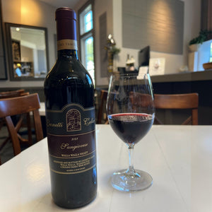 A bottle of LEONETTI SANGIOVESE 2023 by WINE: WINEBOW rests on a countertop, while shelves filled with assorted wine bottles appear blurred in the background.