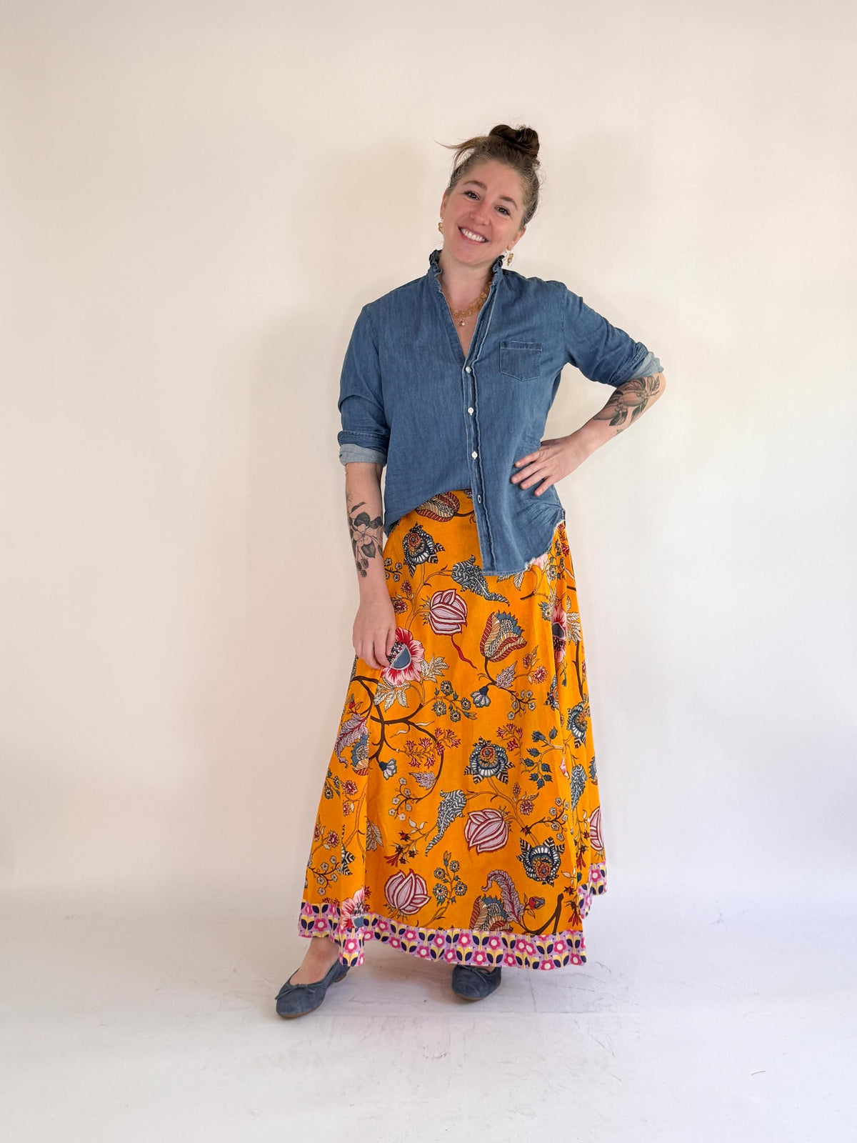 A smiling, tattooed person wears a blue denim shirt, gray shoes, and the FARYLINA MAPLE WRAP SKIRT by FARYLINA—a bright orange floral maxi skirt with pink trim—standing against a plain light background.