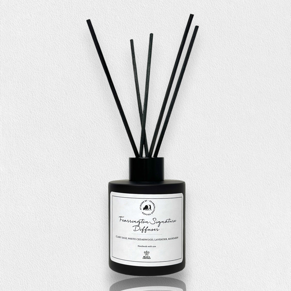 A matte black glass bottle containing several thin black reeds makes an ideal holiday gift. The label on the front reads &quot;Fearrington Signature Diffuser - Clary Sage, White Cedarwood, Calming Lavender, Marjoram.&quot; The backdrop is a white, textured surface.