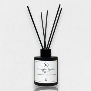 A matte black glass bottle containing several thin black reeds makes an ideal holiday gift. The label on the front reads "Fearrington Signature Diffuser - Clary Sage, White Cedarwood, Calming Lavender, Marjoram." The backdrop is a white, textured surface.
