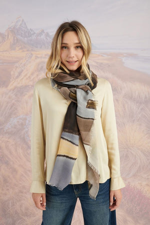 A woman in a black long-sleeve top and beige pants stands before a scenic backdrop, her look elevated by the STORIATIPIC ISCHIA Modal and Wool Scarf (100x200 cm) in vibrant orange and brown, draped elegantly around her neck.