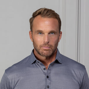 A man with short brown hair and a beard wears the COLLARS AND CO Semi Spread Collar Original Polo in Brown Houndstooth, standing in an urban setting with blurred buildings and warm outdoor lighting in the background.