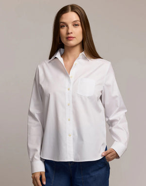 A woman with long brown hair wears the HINSON WU GWEN Long Sleeve No Iron Cotton Shirt in white, featuring a chest pocket, paired with blue jeans. She stands against a light background with one hand in her pocket and a neutral expression.