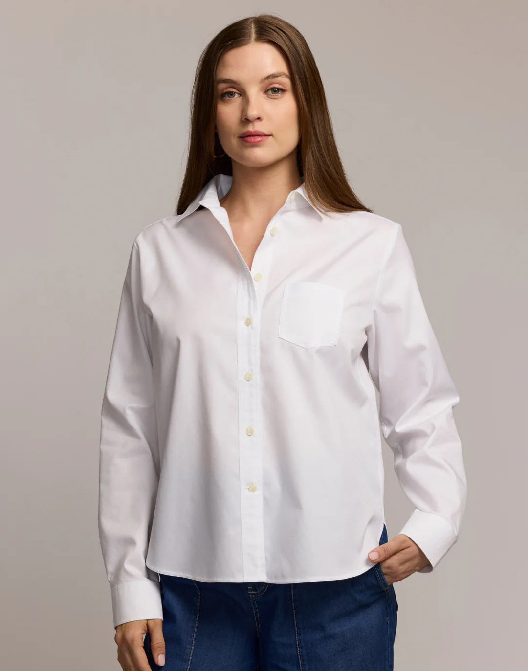 A woman with long brown hair wears the HINSON WU GWEN Long Sleeve No Iron Cotton Shirt in white, featuring a chest pocket, paired with blue jeans. She stands against a light background with one hand in her pocket and a neutral expression.