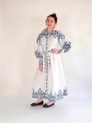 A woman with light brown hair stands on a paved sidewalk in front of a cream-colored building, wearing the FARYLINA SHERI LONG SLEEVE DRESS by FARYLINA—white cotton with blue florals, indigo embroidery, and a belted waist—plus black shoes.