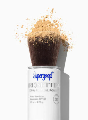 A container of SUPERGOOP 100% MINERAL POWDER SPF35 with built-in brush sits on a white surface, surrounded by scattered light brown powder. This SUPERGOOP powder sets makeup and controls shine.