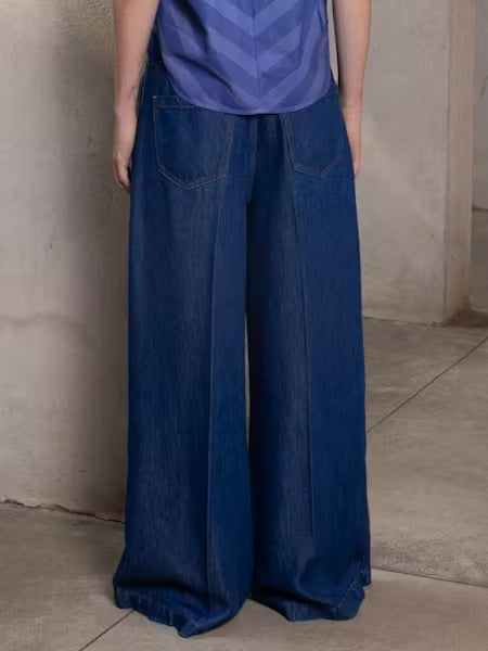 A person stands indoors, shown from the waist down, wearing NUDE's TILDA WIDE LEG DENIM TROUSER with an ultra wide-leg silhouette covering their shoes, set against beige walls and a tiled floor.