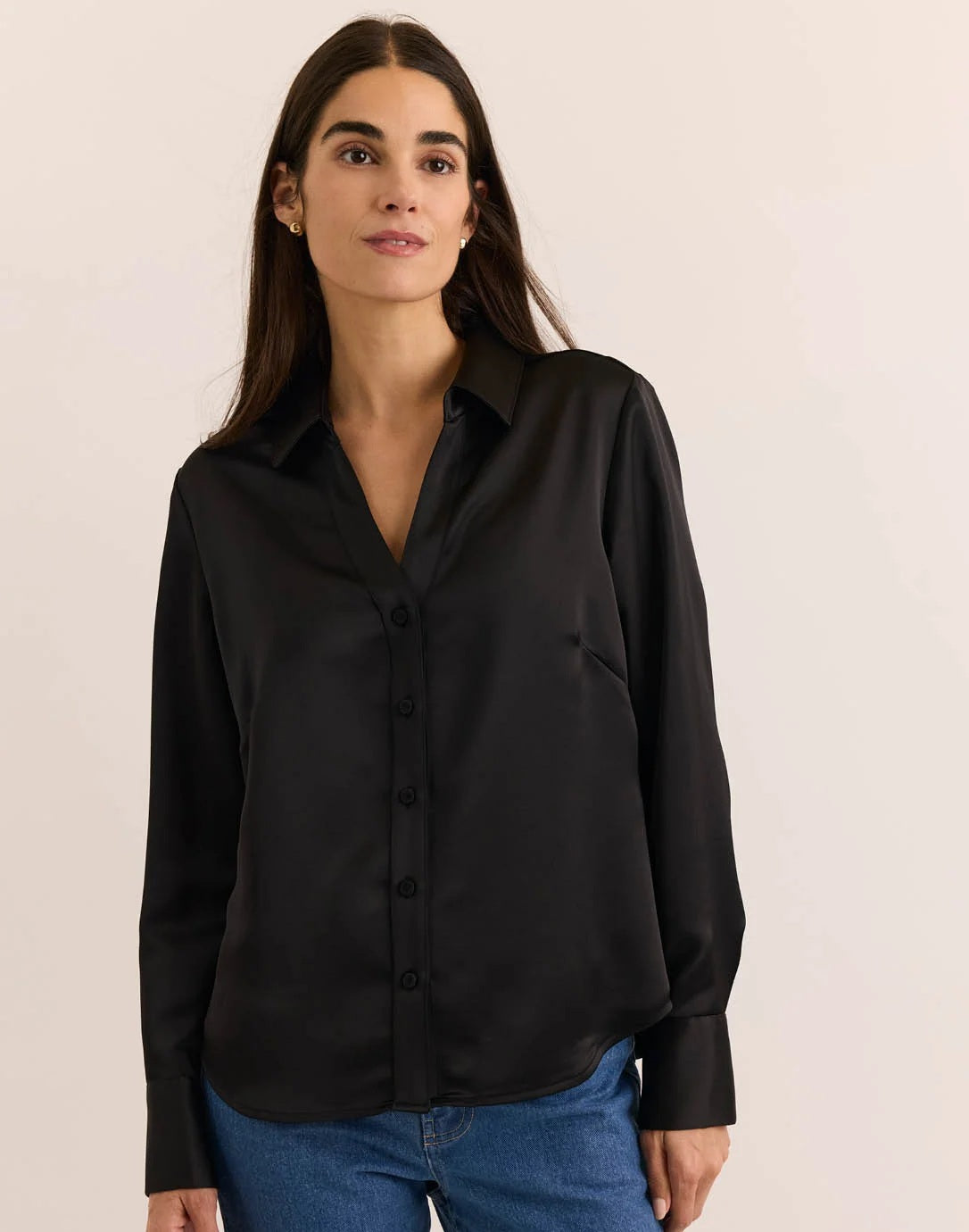 A woman with long dark hair is wearing the HINSON WU Lindsey Long Sleeve Liquid Satin Shirt in Black, which features a relaxed fit, paired with blue jeans. She stands against a plain light-colored background, looking at the camera with a slight smile.