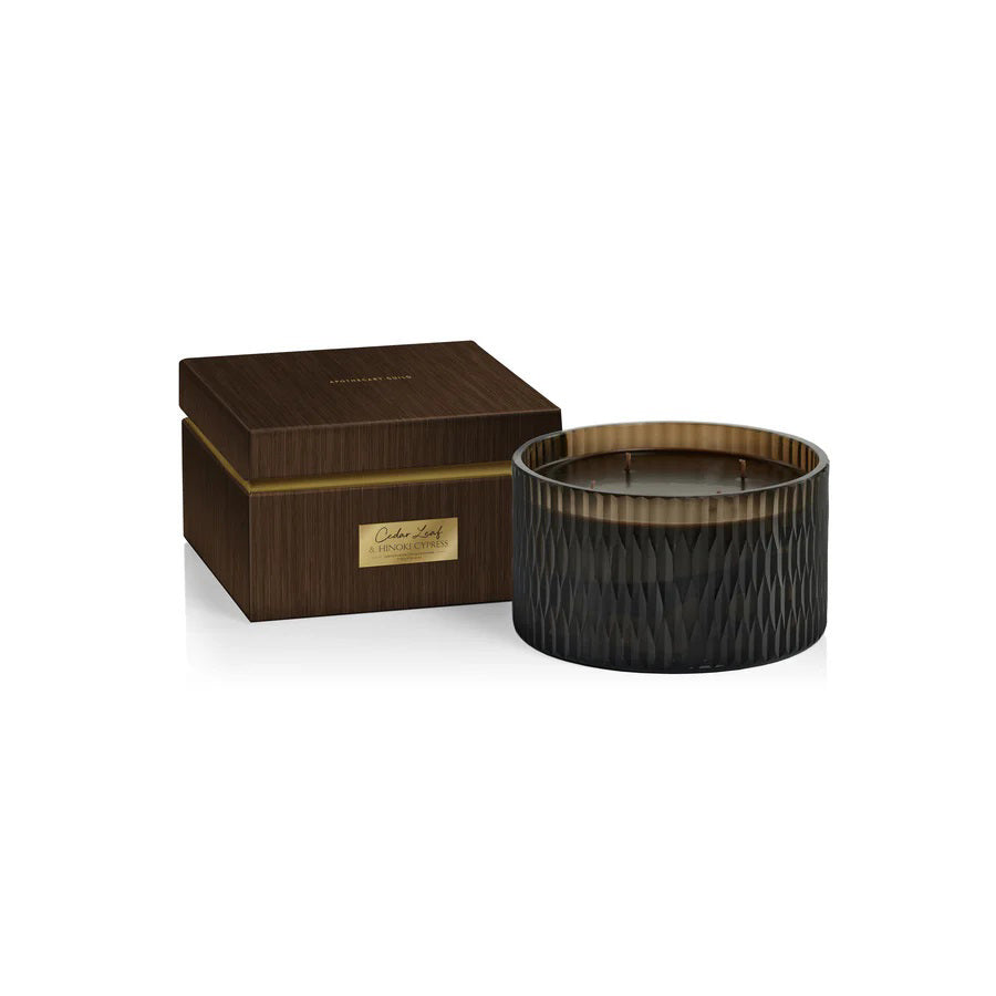 A cylindrical, dark green candle with a textured surface sits next to a matching dark brown, wood-textured box with a gold label. The box lid reads &quot;ZODAX.&quot; The candle features five wicks and boasts fragrance notes of Cedar Leaf and Hinoki Cypress, with the box label reading &quot;5 WICK CANDLE - CEDAR LEAF &amp; HINOKI CYPRESS&quot; to indicate the scent.