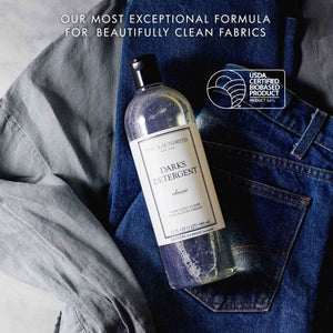 A clear bottle of THE LAUNDRESS - DARKS DETERGENT 32OZ with a black cap is displayed against a white background. The label reads "Darks Detergent Classic, formulated with an enzyme blend to keep dark laundry vibrant, 32 fl oz (1 qt) 946 ml." A caution note is visible at the bottom.