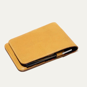 The WILL LEATHER GOODS - GOLF YARDAGE BOOK IN YELLOW by WILL LEATHER GOODS is a yellow leather case holding a spiral-bound yardage book with nature illustrations, shown open with a pen beside it on a white background.
