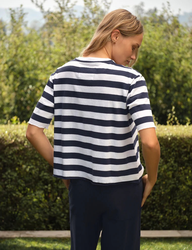 A woman stands outdoors in front of greenery, wearing the FRANK & EILEEN PERRY NYC Crewneck Tee in Vintage Jersey Nautical Stripe and dark pants, with her hands in her pockets, smiling slightly at the camera.