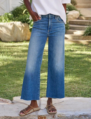 A person stands outdoors on grass, wearing the FRANK AND EILEEN - GALWAY WIDE LEG JEAN IN 2002 MID WASH and a white shirt, with their right hand relaxed by their side.