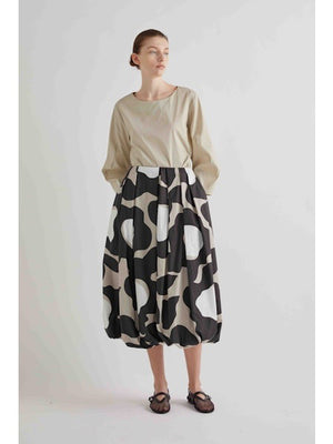A person wearing a beige long-sleeve top, the MEIMEIJ BLACK MOONBEAM SKIRT—a voluminous cotton midi with a bold black, beige, and white abstract print—and black mesh sandals stands against a plain light background.