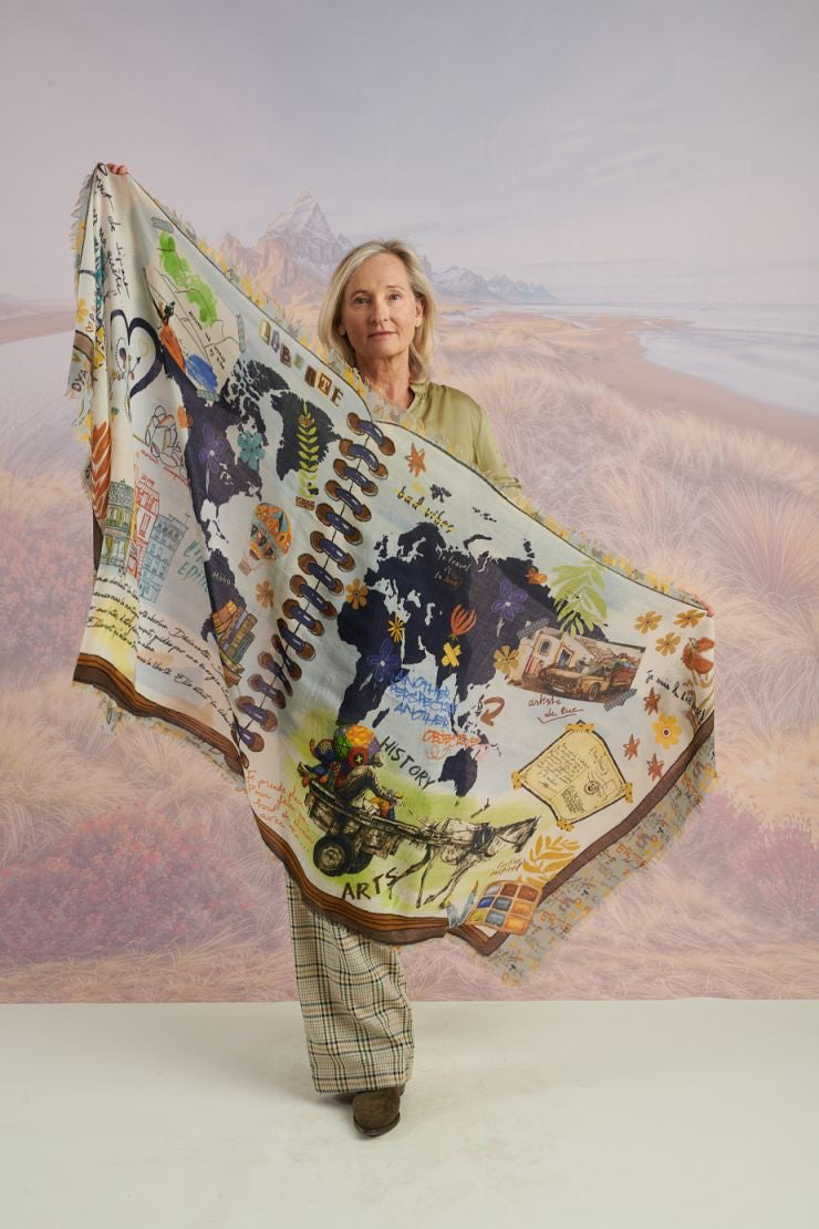 A woman with wavy blonde hair smiles while looking down. She wears a loose, light lavender blouse, dark pants, and the STORIATIPIC EUGENIE WOOL SCARF. Behind her is a scenic landscape of mountains, grass, and water.