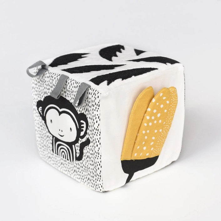 The WEE GALLERY - JUNGLE SOFT BLOCK by WEE GALLERY is a soft fabric cube perfect for stimulating a baby's developing senses. It features monochrome patterns and playful designs, including a cute monkey illustration on one side and a yellow leaf on another. Inspired by jungle life and animals, this toy boasts various textures and small loops on top for easy grasping.
