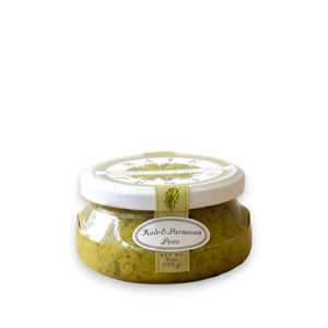 A small, round jar with a white lid labeled "BELLA CUCINA - KALE & PARMESAN PESTO." The jar is filled with green, all-natural pesto sauce and features a decorative label displaying the product name and net weight of 6 ounces (169 grams).