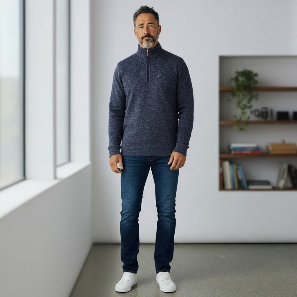 A man with short dark hair and a beard wears the CRIQUET - CROSSROADS Pullover in Moody Blue from CRIQUET over a white shirt and blue jeans. He stands facing forward against a plain white background.